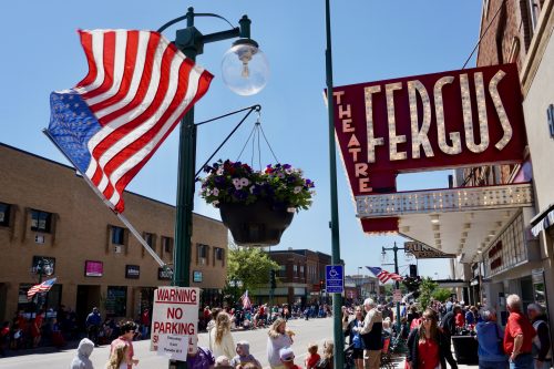 A crowd of people line the street in downtown Fergus Falls in anticipation of a parade. An American flag flaps in the wind and the marquee of the Fergus Theater is lit up.